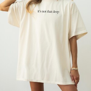 May include: An oversized white t-shirt with the text "it's not that deep" printed on the front. The shirt is made from a soft, comfortable fabric and has a relaxed fit.