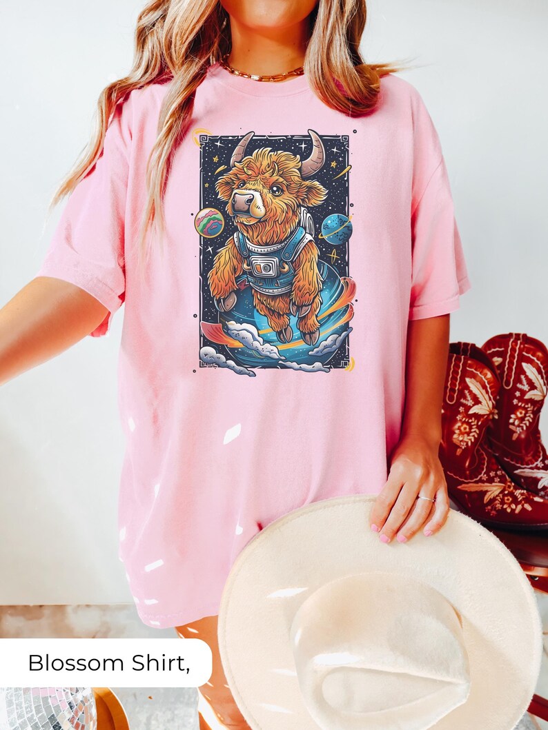 May include: A pink t-shirt with a white graphic print of a brown highland cow wearing a space suit and floating in space. The cow is surrounded by stars and planets. The text "Blossom Shirt" is printed below the image.