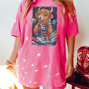 May include: A pink oversized t-shirt with a graphic of a brown and white highland cow wearing a spacesuit and floating in space. The cow is surrounded by stars and planets.