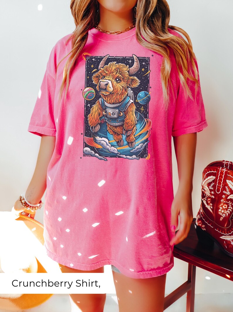 May include: A pink oversized t-shirt with a graphic of a brown and white highland cow wearing a spacesuit and floating in space. The cow is surrounded by stars and planets.
