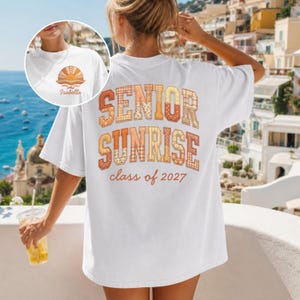 以下が含まれることがあります： 背面にオレンジ、黄色、茶色の模様の文字で「SENIOR SUNRISE class of 2027」と書かれた白いTシャツ。前面には「Isabella」という名前の夕日のデザインが描かれた小さな画像があります。