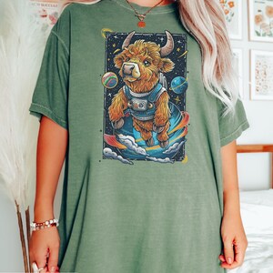 May include: A moss green oversized t-shirt with a graphic print of a cartoon highland cow wearing a space suit and floating in space. The cow is brown and white with a black nose and hooves. The background is black with stars and planets.