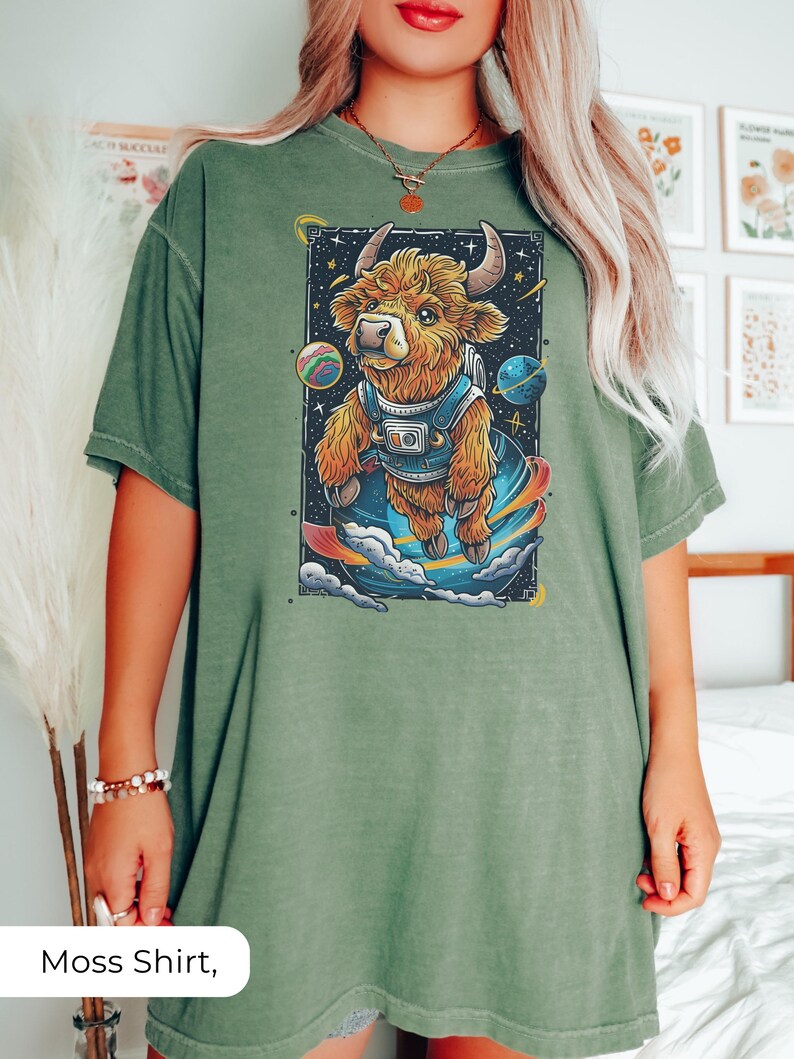 May include: A moss green oversized t-shirt with a graphic print of a cartoon highland cow wearing a space suit and floating in space. The cow is brown and white with a black nose and hooves. The background is black with stars and planets.