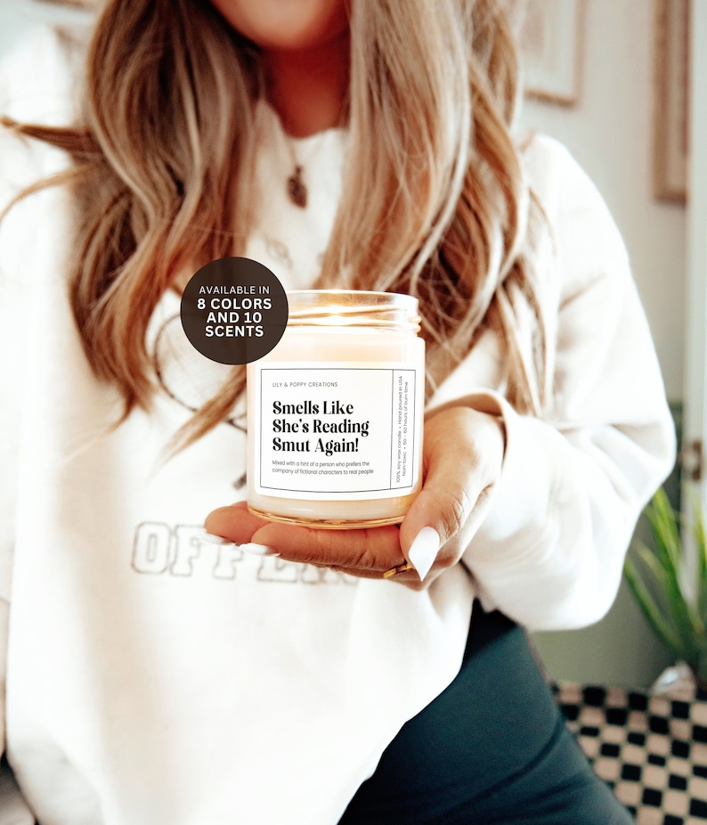 Puede incluir: Una vela blanca con una tapa dorada y una etiqueta que dice "Smells Like She's Reading Smut Again!" La vela est&aacute; en la mano de una persona que lleva una sudadera blanca con la palabra "OFF" en ella. Disponible en 8 colores y 10 aromas.