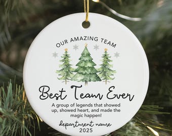 May include: A white, round Christmas ornament with a gold string. The ornament features the text "OUR AMAZING TEAM" and "Best Team Ever" with a watercolor illustration of three green Christmas trees. The text "A group of legends..." is also included.