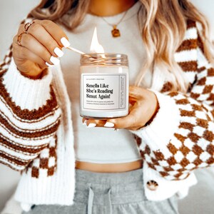 Puede incluir: Una vela blanca con una etiqueta negra que dice "Smells Like She's Reading Smut Again!" La vela se est&aacute; encendiendo con una cerilla.