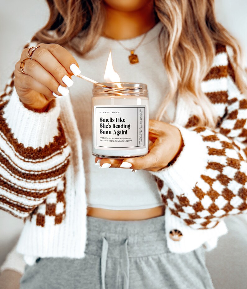 Puede incluir: Una vela blanca con una etiqueta negra que dice "Smells Like She's Reading Smut Again!" La vela se est&aacute; encendiendo con una cerilla.