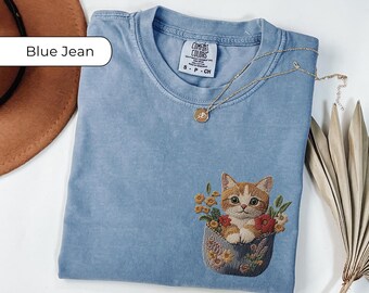 May include: A blue jean t-shirt with a pocket featuring an embroidered orange tabby cat with flowers. The text "COMFORT COLORS" is visible on the shirt.
