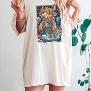 May include: A woman wearing an ivory oversized t-shirt with a graphic of a cartoon highland cow wearing a spacesuit and floating in space. The cow is surrounded by planets and stars.