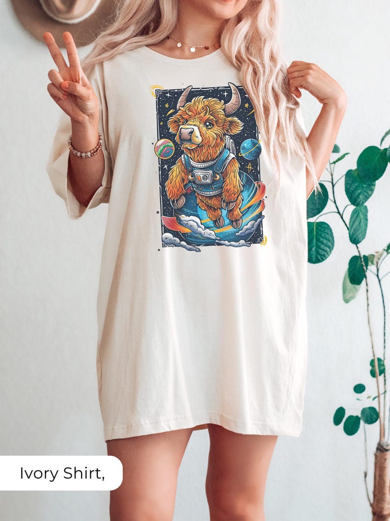 May include: A woman wearing an ivory oversized t-shirt with a graphic of a cartoon highland cow wearing a spacesuit and floating in space. The cow is surrounded by planets and stars.