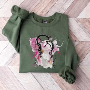 May include: A green sweatshirt with a graphic print of a cat wearing headphones. The cat is white with pink ears and a pink nose. The background of the graphic is a watercolor-style design with pink, blue, and yellow colors.