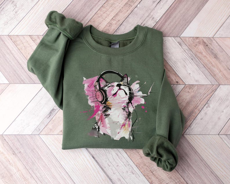 May include: A green sweatshirt with a graphic print of a cat wearing headphones. The cat is white with pink ears and a pink nose. The background of the graphic is a watercolor-style design with pink, blue, and yellow colors.