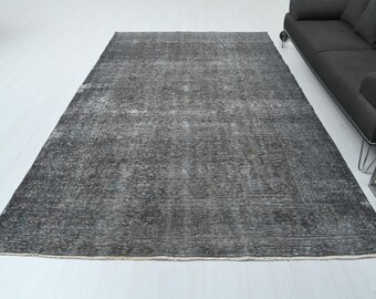 May include: A large, rectangular, distressed-look rug in shades of gray and charcoal. The rug has a faded, vintage appearance with a subtle pattern. It is placed on a white surface, with a dark gray sofa visible in the background.