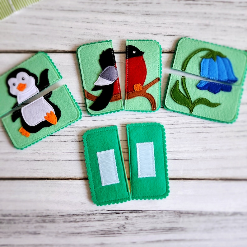 May include: A set of felt puzzle pieces featuring various animals and a flower. The pieces are green with a scalloped edge and have different images on them, including a penguin, a bird, a red bird, and a blue flower.