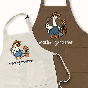 May include: Two aprons, one cream and one brown, with gardening-themed illustrations. The cream apron says "mini gardener" and the brown apron says "master gardener". Each apron features a cartoon goose wearing overalls and a straw hat, holding gardening tools.
