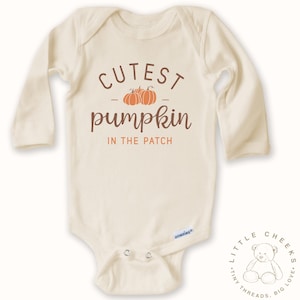 May include: Cream-colored baby onesie with long sleeves. The front features the text "Cutest Pumpkin in the Patch" in brown script and a pumpkin graphic. A small logo is visible near the bottom. The onesie is designed for infants.