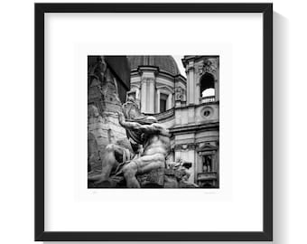 May include: Black and white photograph of a classical sculpture in a square black frame. The sculpture depicts a muscular figure with a draped cloth, set against a backdrop of a building with a dome and architectural details.