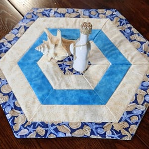 May include: A hexagonal table runner with a coastal theme. The runner features a blue and beige geometric pattern, with a border of seashells and starfish. A white bottle and a seashell are placed on top.