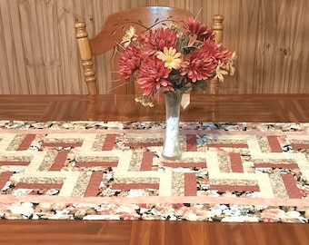 May include: A rectangular table runner with a geometric pattern in shades of red, beige, and floral prints. A vase with red and yellow flowers sits on the runner. The runner is approximately 60 inches long.