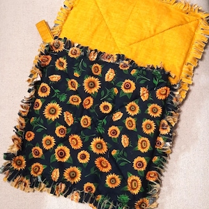 May include: Two square pot holders. One is solid yellow with a loop for hanging. The other is black with a sunflower pattern and a fringed edge. The pot holders are made of fabric.