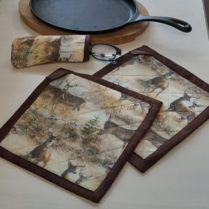 May include: Two brown quilted pot holders with a deer print design. The pot holders have a brown border. A black cast iron skillet is on a wooden cutting board. A pair of glasses and a brown and white eyeglass case with a deer print design are on the table.