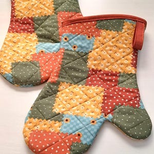 May include: A pair of quilted oven mitts with a patchwork design. The mitts feature a mix of floral and dotted patterns in orange, yellow, blue, and green. The edges are trimmed with a solid orange fabric and have a loop for hanging.