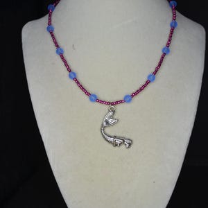 May include: A necklace featuring a silver-toned mermaid tail charm suspended from a strand of small, metallic magenta seed beads interspersed with larger, round, translucent light blue beads, creating a colorful and detailed accessory.