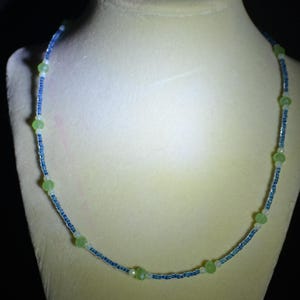 May include: A delicate necklace featuring small blue and white beads interspersed with larger, light green beads. The necklace is displayed on a mannequin, showcasing its design and color palette. The beads are strung together, creating a subtle, elegant accessory.