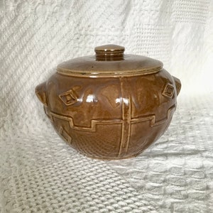 May include: A brown ceramic lidded pot with a decorative geometric design. The pot has two small handles and a round knob on the lid. The pot is sitting on a white textured surface.