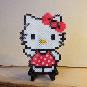 May include: A white Hello Kitty character with a red bow and a red and white polka dot dress, made with perler beads.