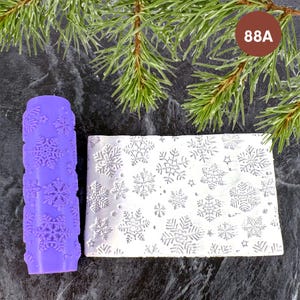 Puede incluir: Un rodillo cilíndrico morado con relieve y una superficie plana rectangular blanca, ambos con un diseño de copos de nieve y estrellas. El rodillo está colocado verticalmente junto a la pieza plana que muestra el patrón invernal.