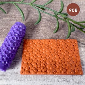 May include: A purple cylindrical rolling pin and a rectangular flat sheet, both featuring a raised floral pattern of daisies. The text 90B is visible in a brown circle in the upper right corner of the image.