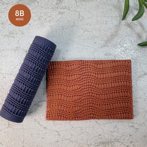 Puede incluir: Un rodillo de textura cilíndrico de color morado oscuro etiquetado como 8B mini, con un patrón geométrico repetitivo de puntos y líneas, junto a una placa rectangular de arcilla color terracota grabada con el mismo patrón intrincado.