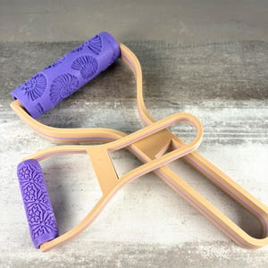 Puede incluir: Una herramienta de rodillo para arcilla de doble extremo con dos cabezales cilíndricos morados con patrones en relieve. El rodillo superior grande muestra un diseño de fósiles de amonita, mientras que el inferior pequeño tiene un motivo floral. Marco beige.