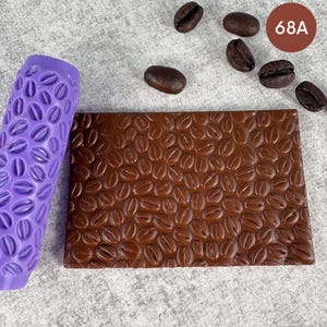 Puede incluir: Un rodillo cilíndrico morado con un patrón de granos de café en relieve junto a una placa rectangular marrón grabada con el mismo diseño. Varios granos de café tostados oscuros están esparcidos cerca y un círculo marrón muestra 68A.