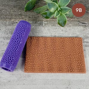 May include: A purple cylindrical patterned roller tool next to a rectangular brown slab featuring an embossed cobblestone texture. The roller has a repeating honeycomb-like design that matches the pattern imprinted on the brown rectangular surface.