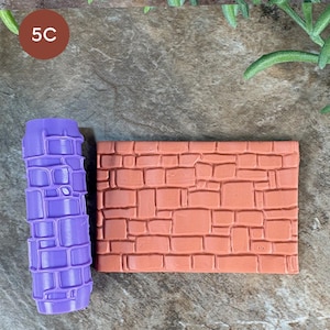 May include: A cylindrical purple roller tool featuring a raised stone wall pattern alongside a rectangular terracotta-coloured clay slab embossed with the same repeating irregular brick design, showing the texture created by the roller.