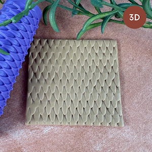 May include: A square piece of tan clay featuring a raised, repeating dragon scale texture. Beside it lies a purple cylindrical rolling pin with the same scale pattern embossed on its surface. A small brown circle with the text 3D is visible.