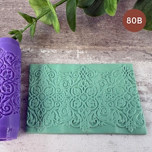 Può includere: Un rullo cilindrico viola e una lastra rettangolare verde, entrambi caratterizzati da un intricato motivo floreale e a volute in rilievo. La lastra verde presenta un design simmetrico ripetitivo e il numero 80B è stampato in un cerchio marrone.