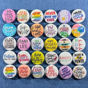 May include: A collection of 35 round pinback buttons with motivational phrases in various colors. The buttons feature phrases like "Dream Big," "Believe in Your Power," and "Do What Makes You Happy."