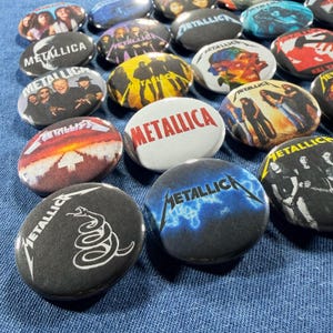 K&ouml;nnte beinhalten: Eine Sammlung runder Metallica-Bandbuttons mit verschiedenen Designs. Einige Buttons zeigen Fotos von Bandmitgliedern, Albumcover und das Logo der Band. Die Buttons sind in den Farbt&ouml;nen Schwarz, Wei&szlig;, Blau, Gelb und Rot gehalten.