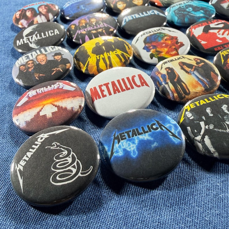 K&ouml;nnte beinhalten: Eine Sammlung runder Metallica-Bandbuttons mit verschiedenen Designs. Einige Buttons zeigen Fotos von Bandmitgliedern, Albumcover und das Logo der Band. Die Buttons sind in den Farbt&ouml;nen Schwarz, Wei&szlig;, Blau, Gelb und Rot gehalten.