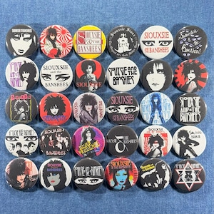 May include: A collection of round Siouxsie and the Banshees buttons. The buttons feature images of Siouxsie Sioux, band logos, and various graphics in black, white, red, yellow, and blue. The buttons are arranged on a denim surface.
