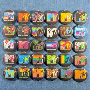 May include: A collection of 28 round MTV pins with various designs. Each pin features the MTV logo and the words "MUSIC TELEVISION." The designs include the American flag, brick patterns, and psychedelic color schemes.