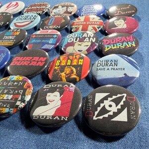 Puede incluir: Una colecci&oacute;n de botones redondos de la banda Duran Duran con varios dise&ntilde;os. Algunos botones presentan el nombre de la banda, retratos o arte de &aacute;lbumes. Los colores incluyen negro, rojo, blanco y azul. Los botones est&aacute;n dispersos sobre una superficie azul.