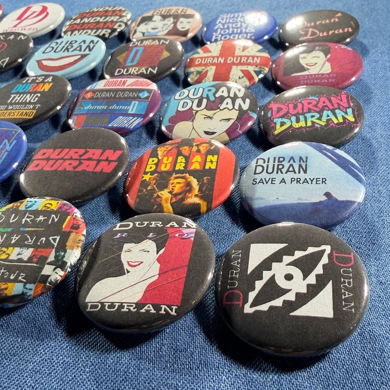 Puede incluir: Una colecci&oacute;n de botones redondos de la banda Duran Duran con varios dise&ntilde;os. Algunos botones presentan el nombre de la banda, retratos o arte de &aacute;lbumes. Los colores incluyen negro, rojo, blanco y azul. Los botones est&aacute;n dispersos sobre una superficie azul.