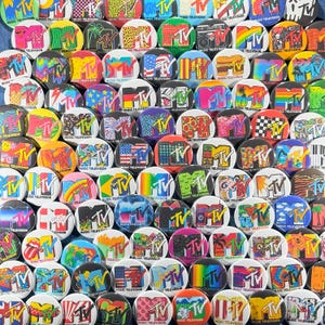 May include: A collection of round MTV Music Television logo buttons. The buttons feature various designs, including the MTV logo, flags, and other graphics. The buttons are in a variety of colors, including red, blue, yellow, and green.