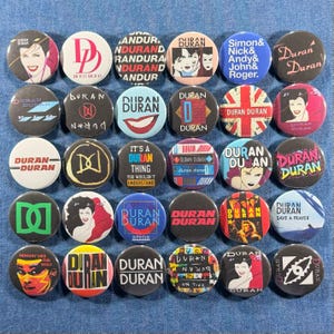 Puede incluir: Colecci&oacute;n de botones redondos de Duran Duran, de aproximadamente 2,5 cm de di&aacute;metro. Los botones presentan varios dise&ntilde;os, incluyendo retratos de miembros de la banda, logotipos y arte de &aacute;lbumes. Los colores incluyen rojo, blanco, negro, azul y amarillo. El texto incluye t&iacute;tulos de canciones y el nombre de la banda.