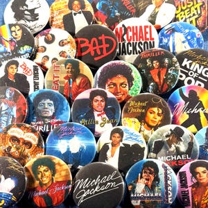 May include: A collection of circular pin-back buttons featuring various photographic portraits, album cover art, and graphic designs of Michael Jackson, including text such as Bad, Thriller, Billie Jean, and King of Pop in diverse colors.