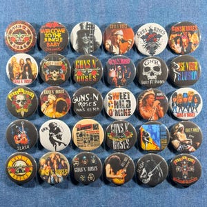 May include: A collection of 30 round pinback buttons featuring various designs related to the rock band Guns N' Roses. The buttons display band members, logos, and song titles in a variety of colors.
